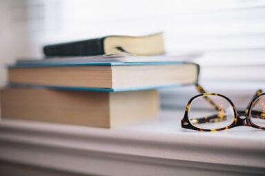 A pair of reading glasses rests on a sunlit window sill next to three books, representing the difference between a psychiatrist, psychologist, and therapist.