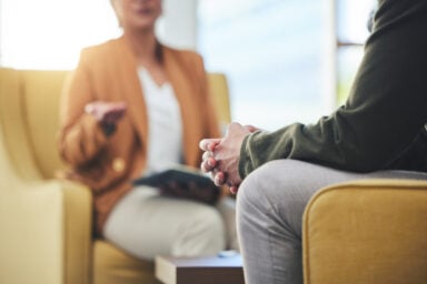 Close-up of a therapy session with two people seated in chairs, one person gesturing while holding a notebook and the other sitting with hands clasped