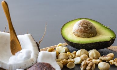 Closeup of section of cutting board with halved avocado, broad wedge of coconut in shell with scoop, macadamia nuts, and walnuts