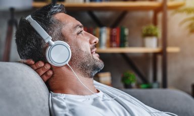Bearded man sits on sofa, reclining into backrest, back of head resting against hand, smiling and relaxed, headphones on