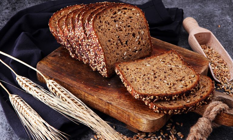 Нарезанный многозерновой хлеб с семенами на разделочной доске, стебли пшеницы с одной стороны, совок с семенами с другой