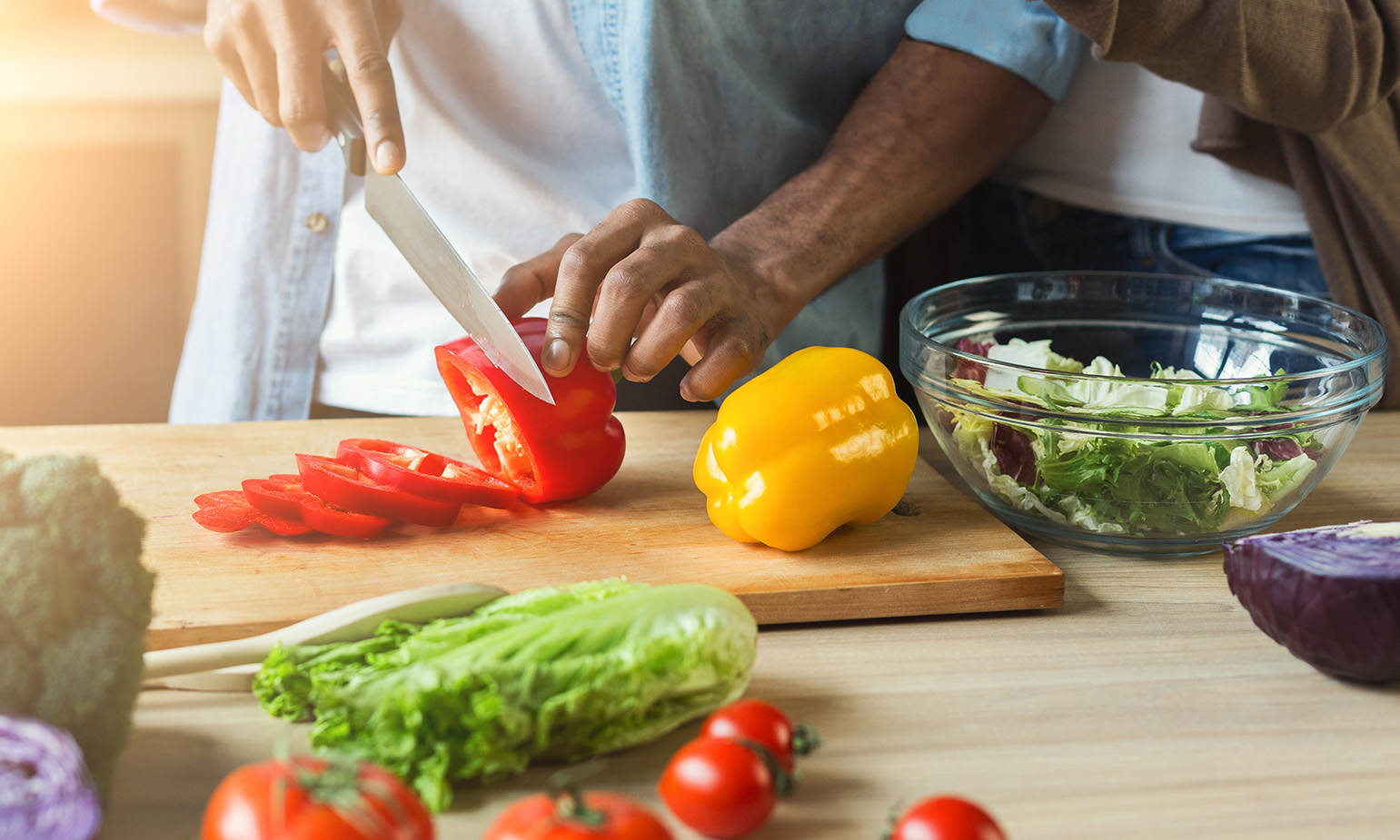 Closeup of bell peppers on cutting board being sliced by man's hands, woman close behind him, other vegetables arrayed on table