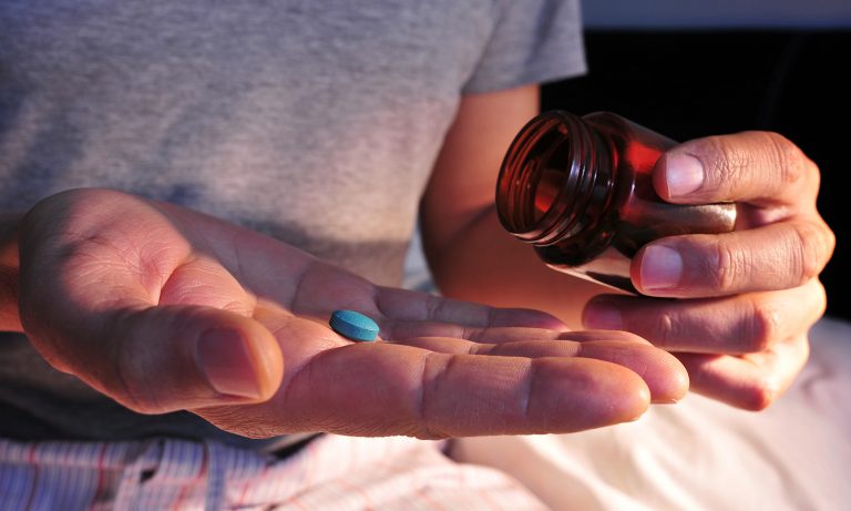 Closeup of man tipping pill bottle grasped in one hand, blue pill in palm of other hand