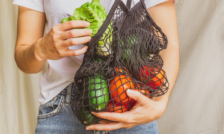 Closeup of mesh satchel full of vegetables slung around a person's shoulders, lettuce in his hand