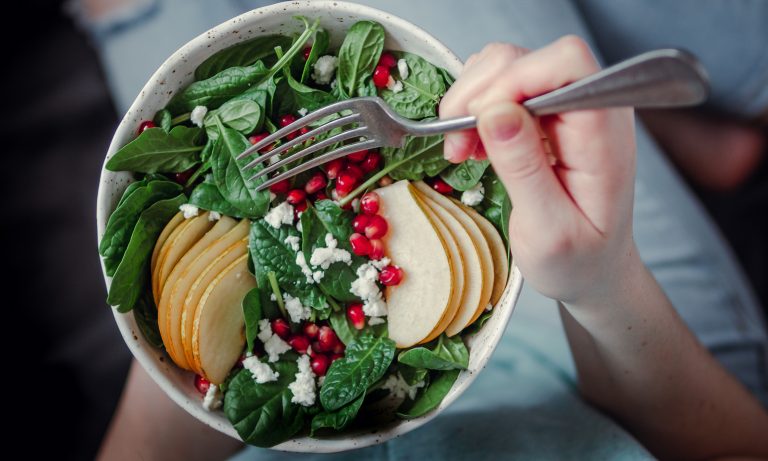 Top-view closeup of salad in bowl, held in hand of girl curled up on sofa, fork at the ready, sliced apples, pomegranate seeds