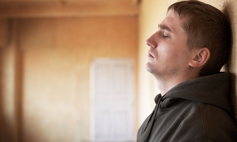 Distressed man standing and leaning against wall of bare-walled room, eyes closed