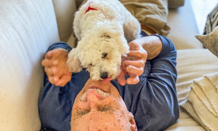 Man reclined on sofa, reacting with gleeful surprise as small dog, on his chest licks his chin