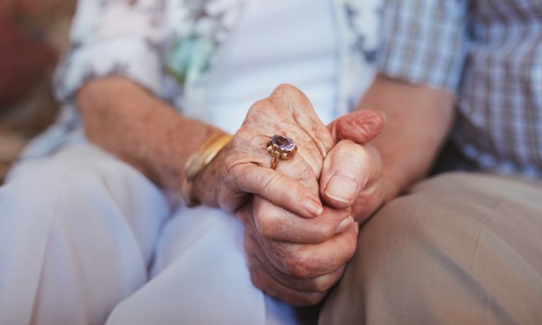 Hands of elderly couple clasped in front of them as they sit side by side