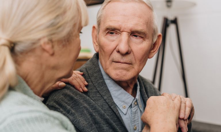 Elderly man, hands clasped over handle of cane, comforted affectionately by his wife