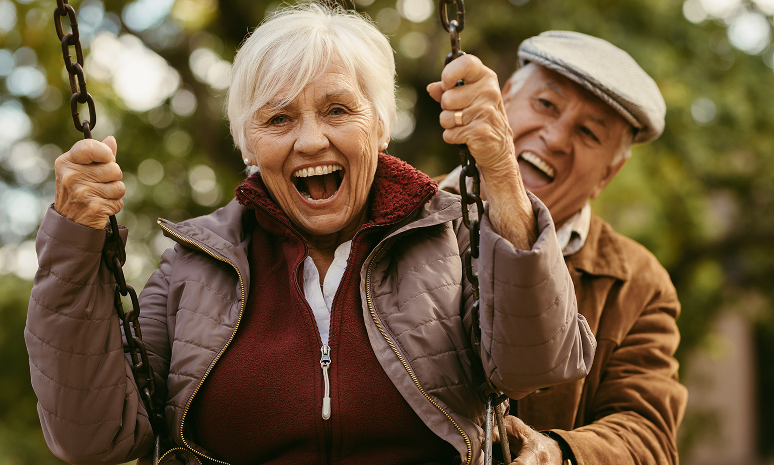 Elderly woman on playground swing, mouth agape in delight as husband gleefully guides swing from behind