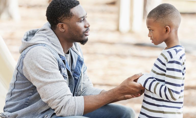Молодой отец сидит и держит за руки своего маленького сына на детской площадке, полностью взаимодействуя с мальчиком, когда он разговаривает с ним.