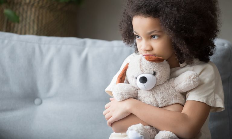 Girl sitting at edge of sofa clutching teddy bear, head tilted down, chin resting on bear's head as she looks forward bashfully