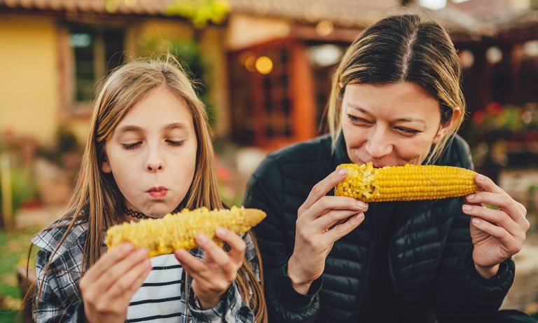 Girl sits in yard beside mother, both eating corn on the cob, the mother smiling as she watches her daughter savor it