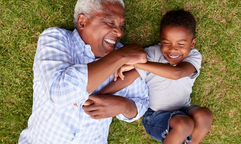 Grandfather play-tussles with young grandson while lying on grass lawn, both smiling></picture><p><a id=