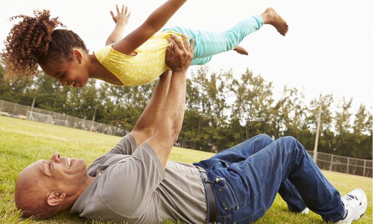 Grandfather and granddaughter smile as grandfather lies on his back in grass field and lifts her into the air above him