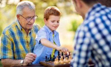 Young boy prepares to grasp a chess piece as he kneels on his grandfather's lap in front of an outdoor chess table