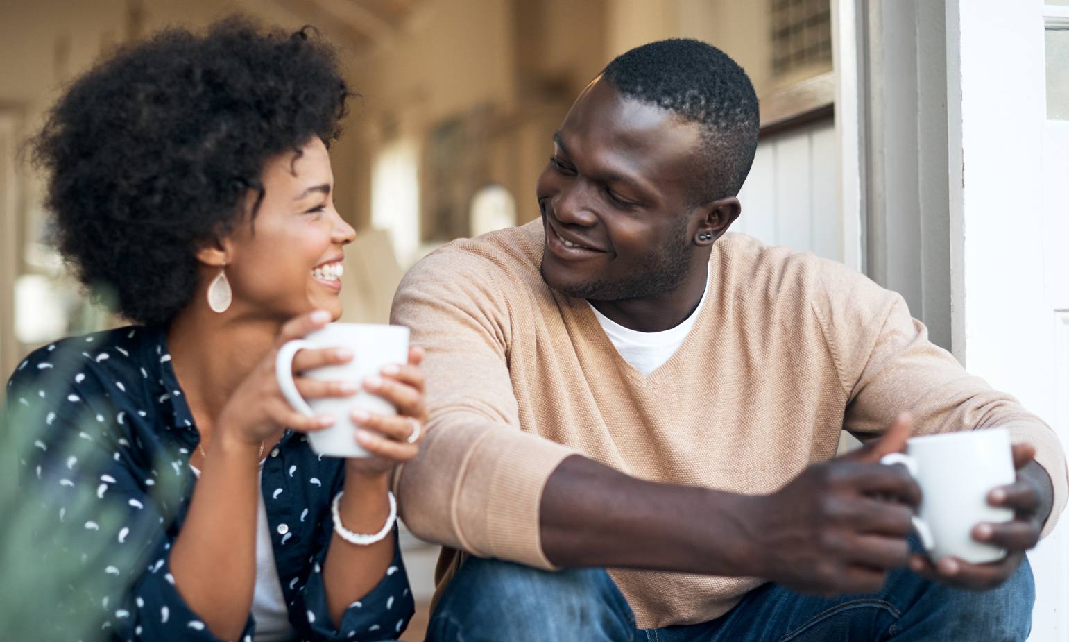 Young woman and man sitting side-by-side on stoop, coffee mugs in hand, looking at each other, smiling