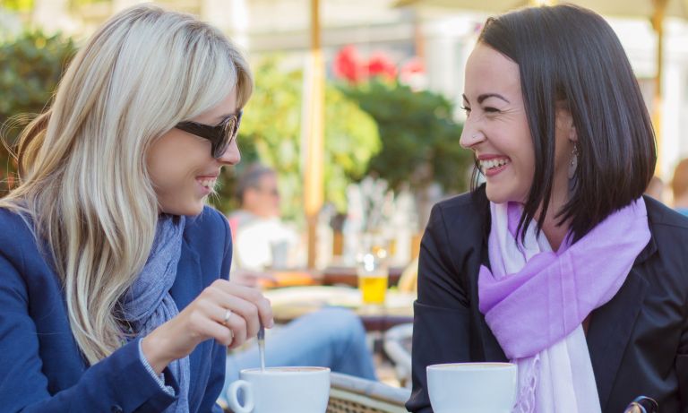 Two women seated together at outdoor cafe table, sharing an intimate laugh