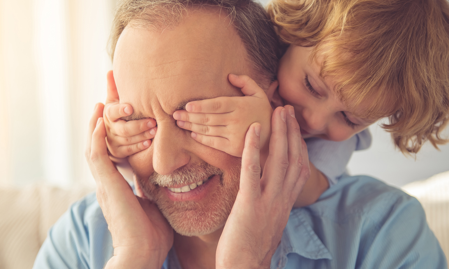 Young child perched on backrest of sofa behind grandpa's head reaches his hands around to cover his grandpa's eyes