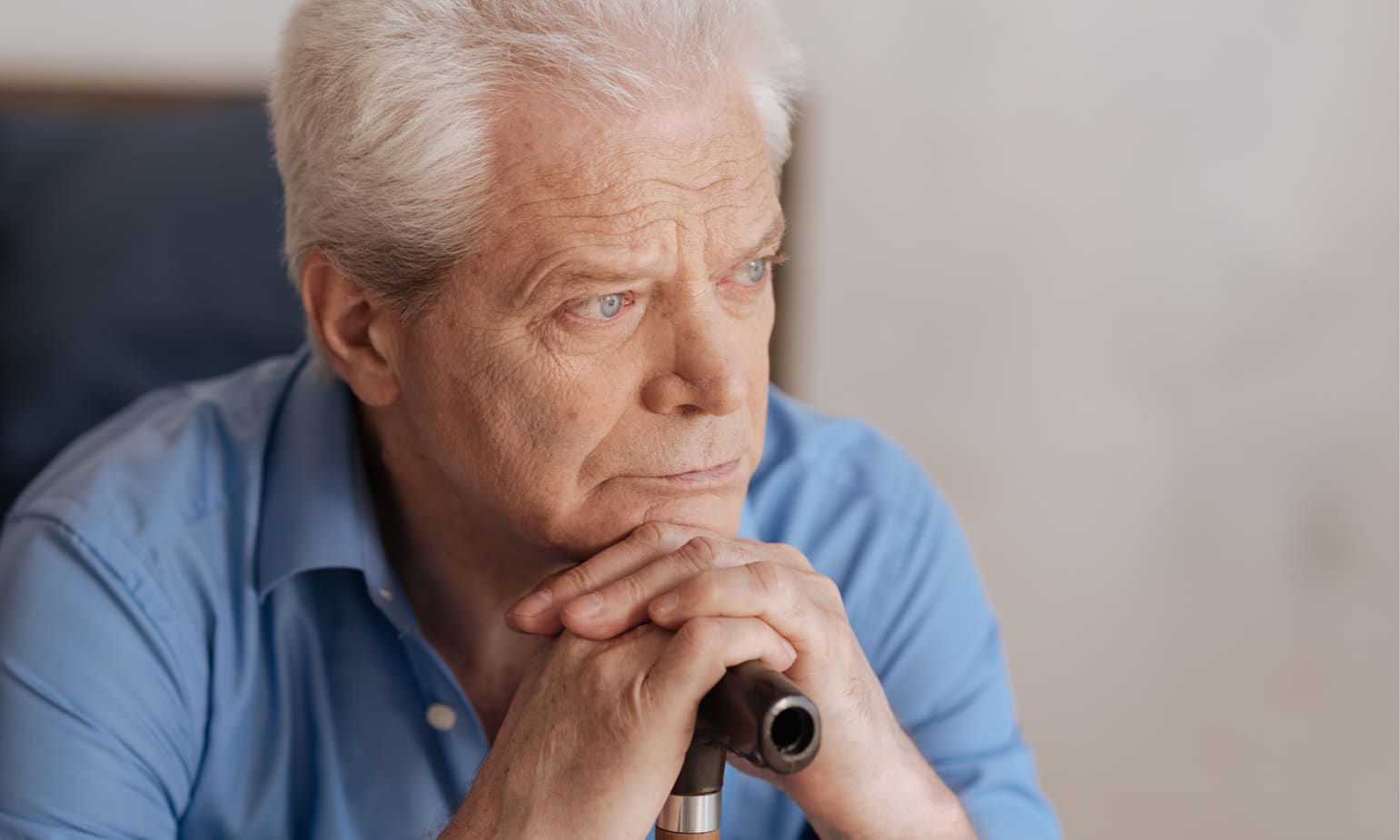 Older man sitting, clasping hands across his cane's handle as he rests his chin there and gazes distantly ahead