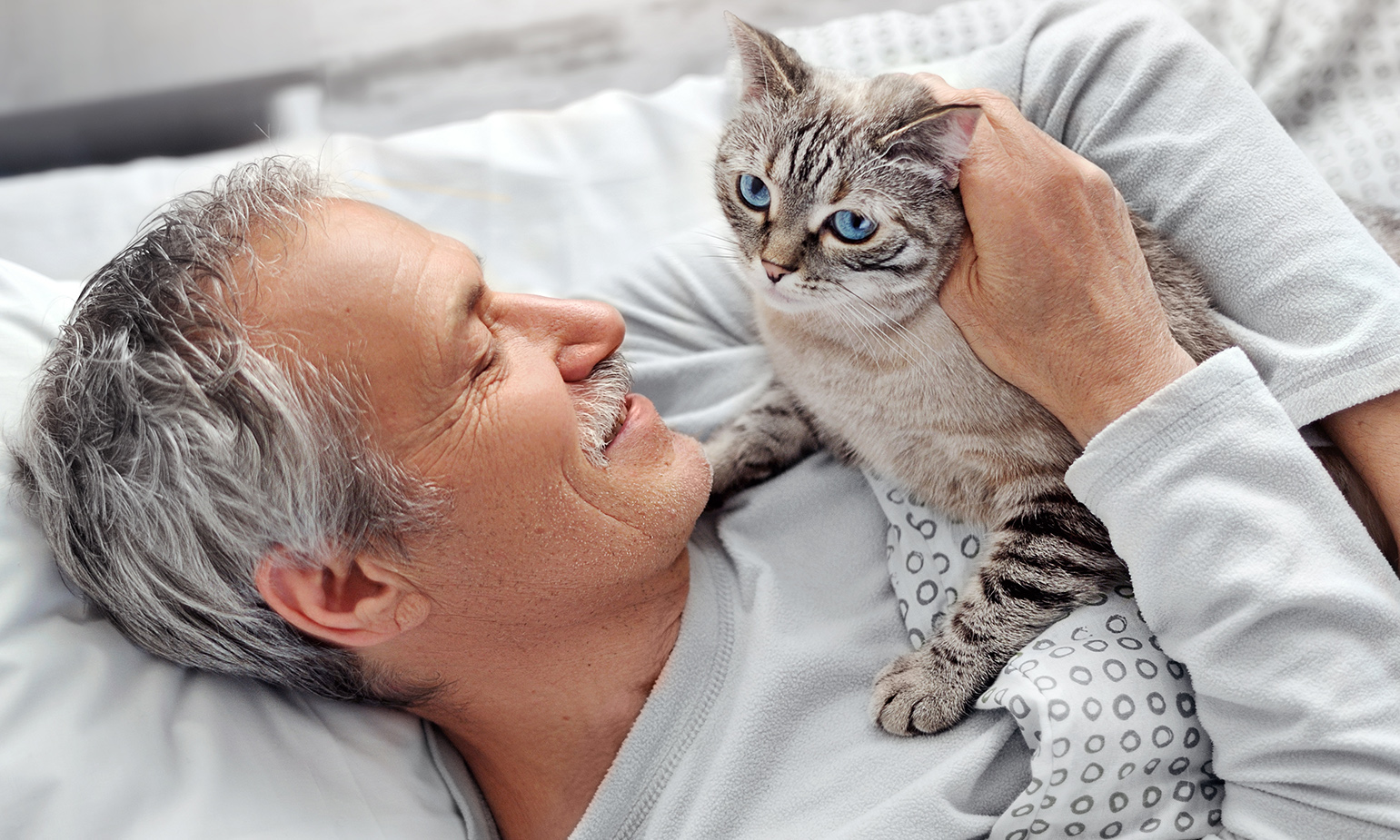 Senior man lying in bed, cradling a cat in his arms and smiling