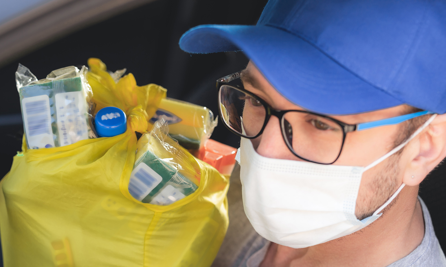 Closeup of man sitting in car's driver's seat, wearing surgical mask and ball cap, holding plastic bag full of groceries