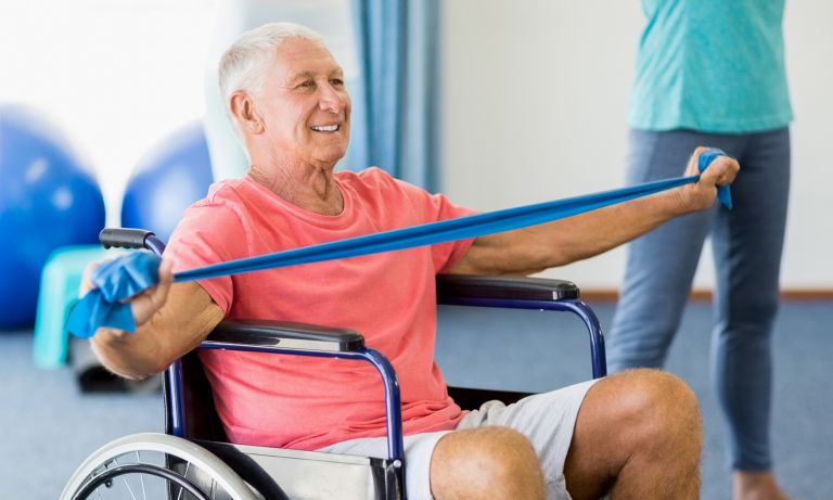 Smiling man seated in wheelchair in group exercise room, pulling a resistance band outward with his arms