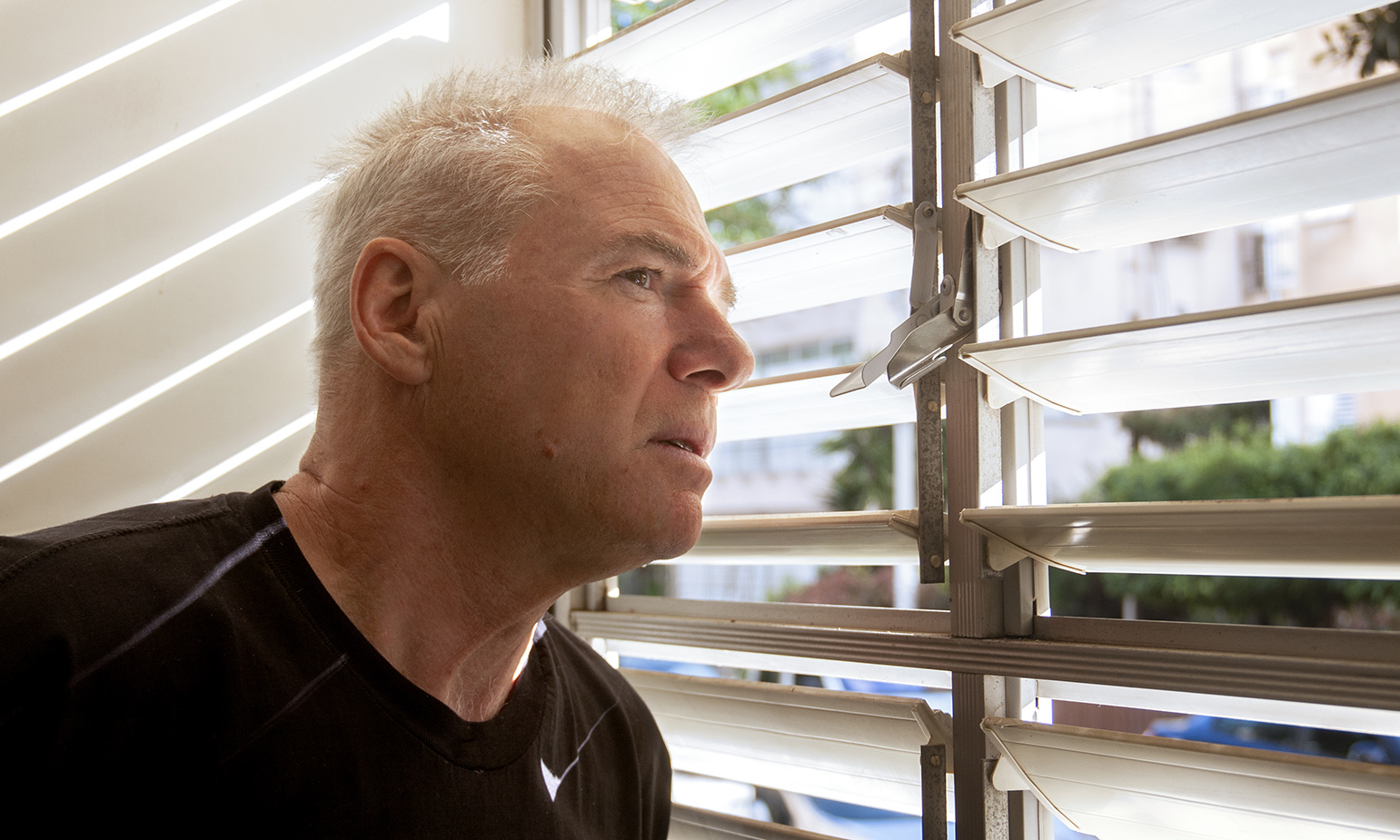 Older man looking out through slats in window blinds towards street warily, his jaw tense