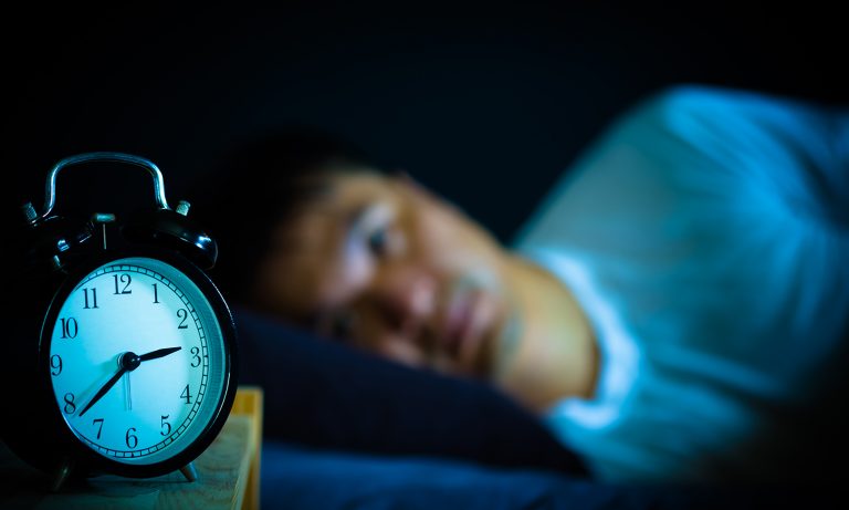 Vyras guli lovoje ir žiūri į žadintuvą pirmame plane, laikrodis rodo 2:40, žadintuvas nustatytas į 6 val.