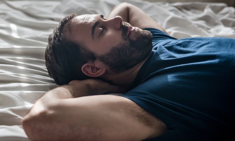 Closeup of young bearded man lying on sheets of bed, hands folded behind bed, looking relaxed