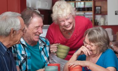 Older friends gathered around kitchen table, clutching ceramic mugs and sharing laughter