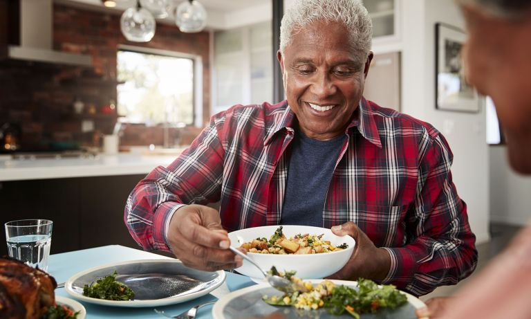 Smiling older man at table in well-appointed kitchen uses serving spoon to fill partner's plate from serving bowl
