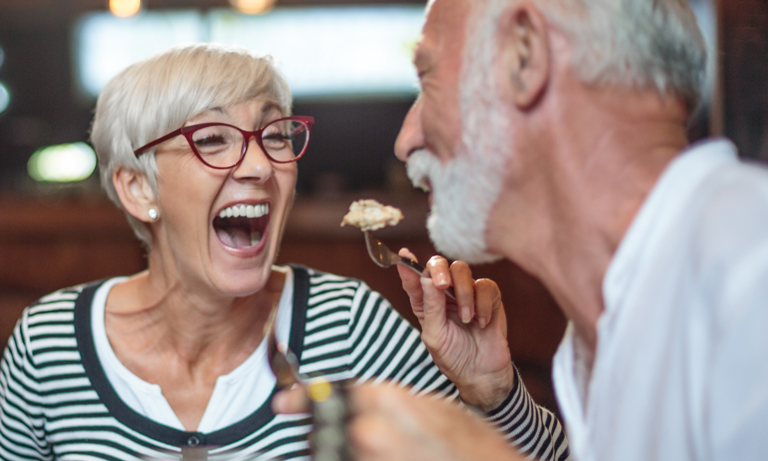 Senior woman laughing while playfully and affectionately feeding her male partner in restaurant