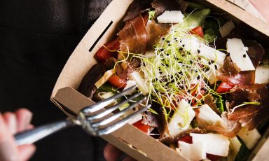 Closeup of salad in takeout box, a fork, grasped in fingers, extending towards it