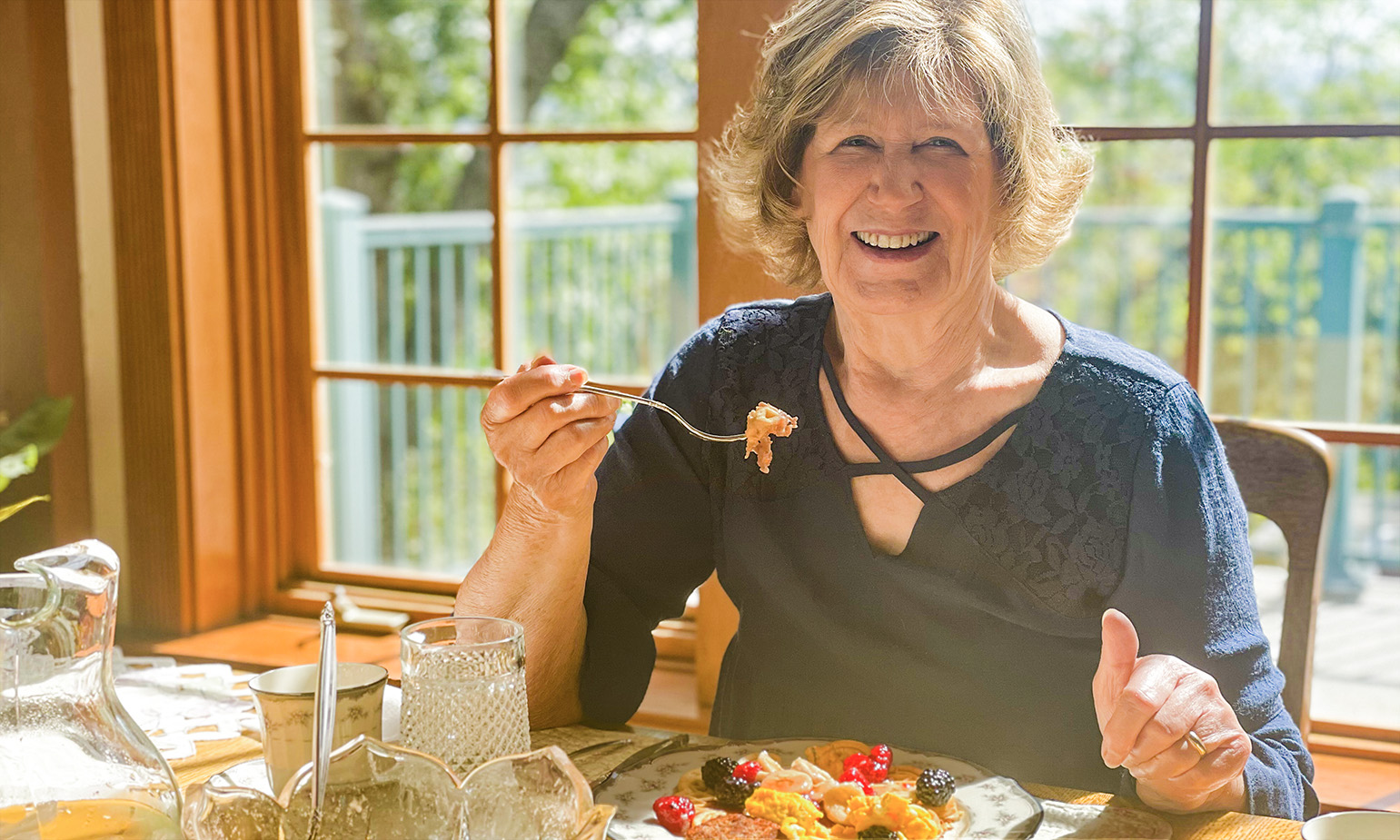 Senior woman enjoying healthy meal at sunlit dining room table at home, picture windows behind her