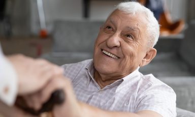 Old man sitting, smiling at person whose hand rests on his as he grips the handle of his cane