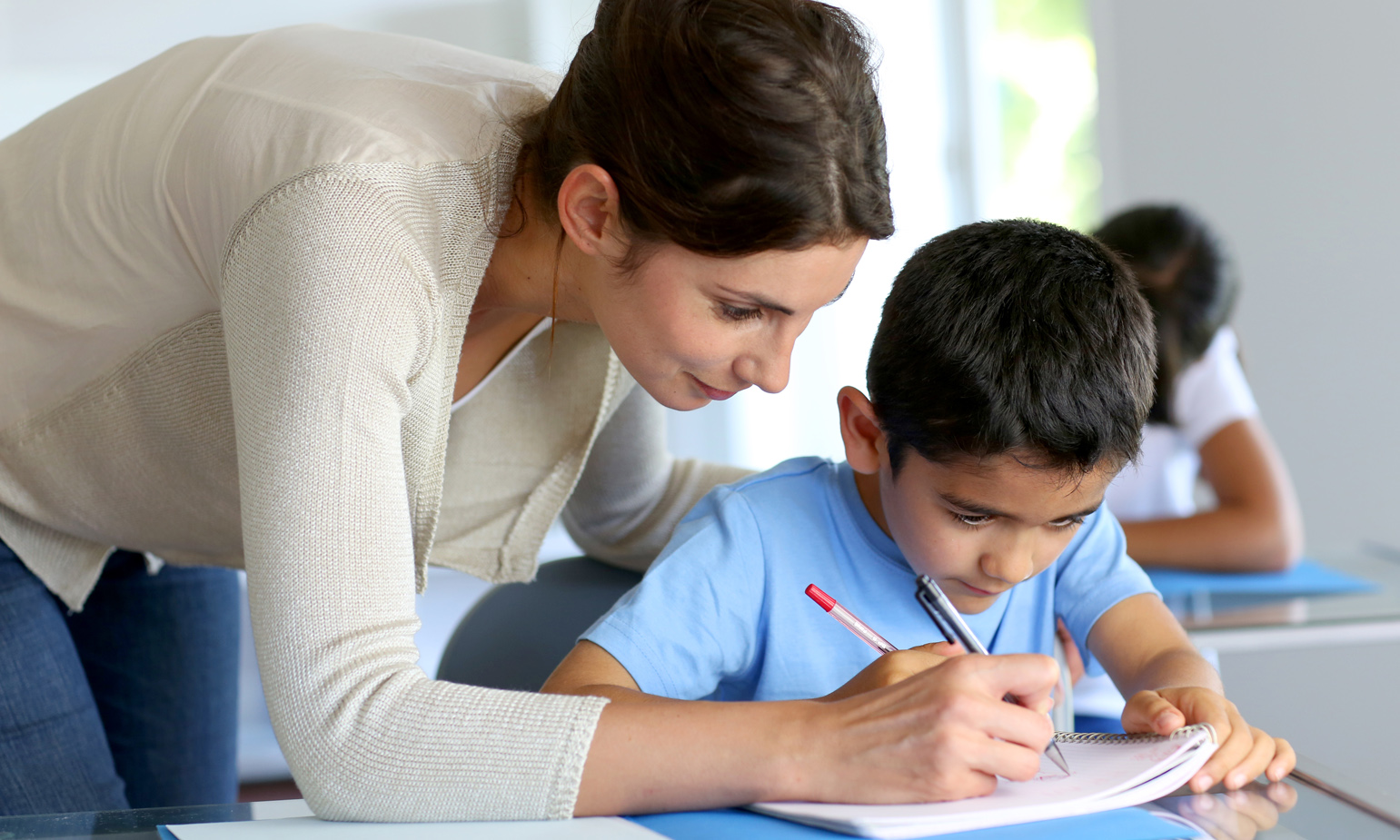 Teacher leaning over shoulder of young boy at his classroom desk, guiding him by marking with her pencil on his notebook