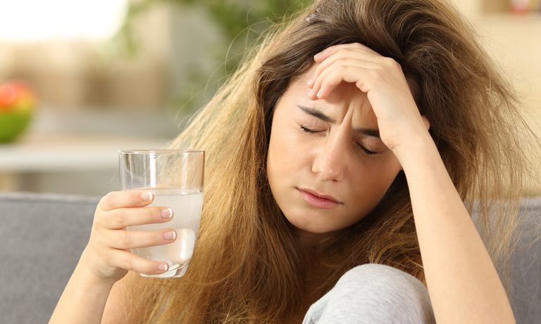Teenage girl holding glass with cold beverage in one hand, other hand on her forehead, eyes closed, pained expression