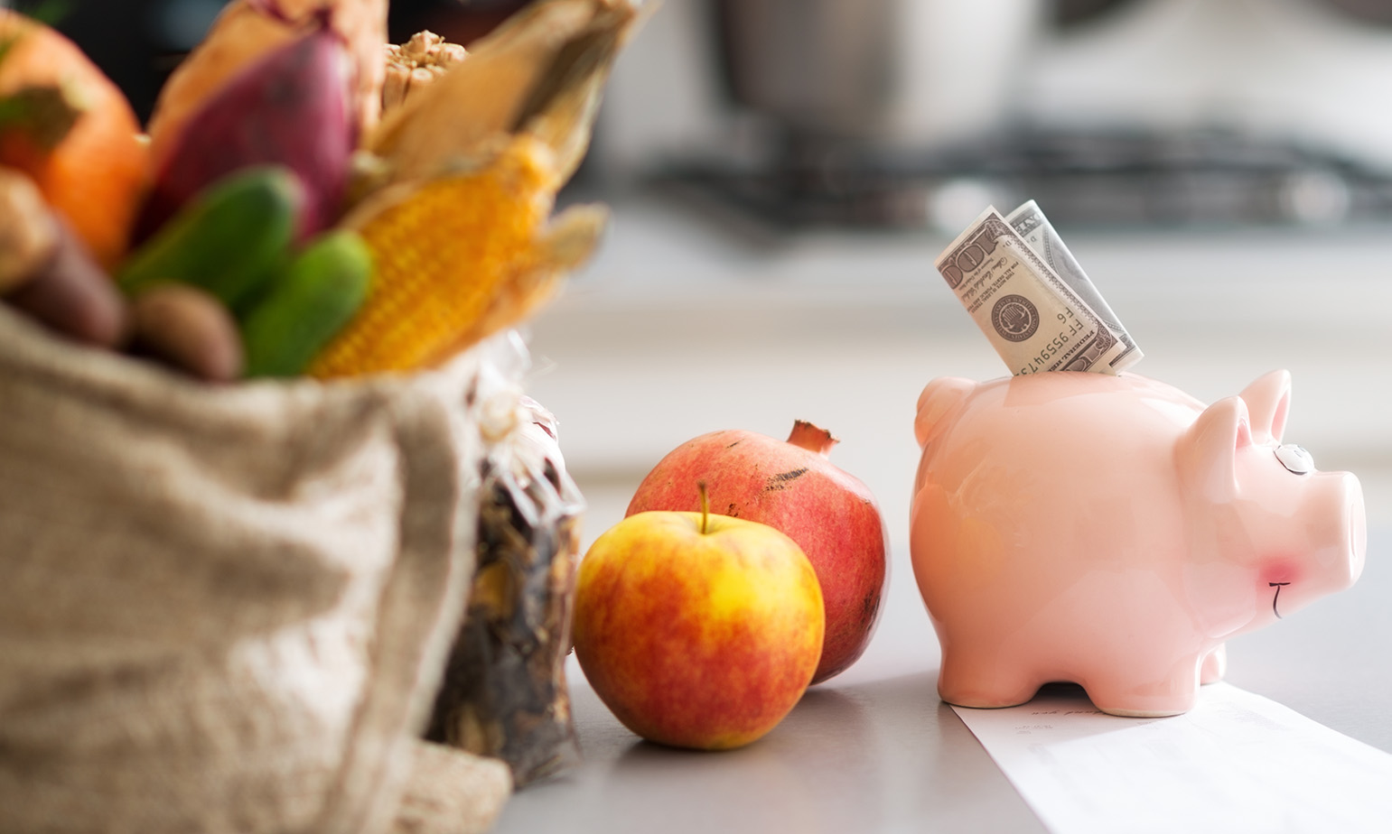 Piggy bank with folded cash bill nestled in slot, in front of bagged produce, pomegranate, and apple, frugal budget food shopping