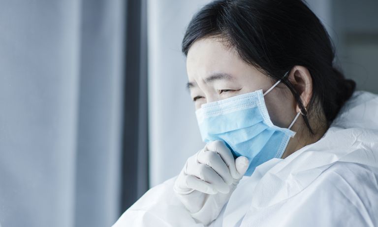 Weary young doctor wearing surgical mask, gloves, and other PPE leans on sill of hospital window looking outward