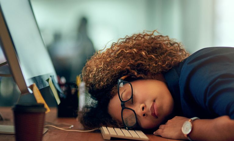 Moteris miega priešais kompiuterį, galva remiasi į klaviatūrą, akiniai kreivai
