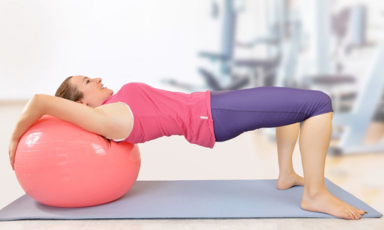 Woman in gym clothes facing upward, leaning back of her shoulders against large exercise ball, elevating torso horizontally