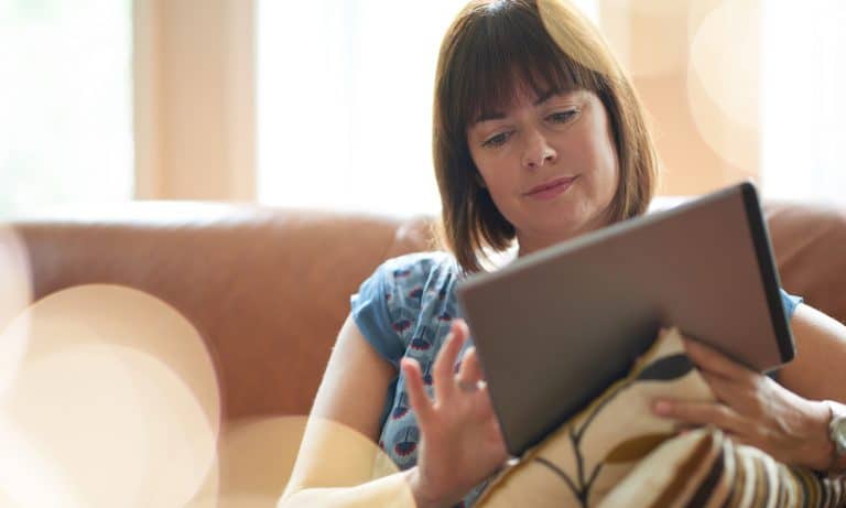 Woman indoors using her tablet, sunlight shining in through translucent white drapes