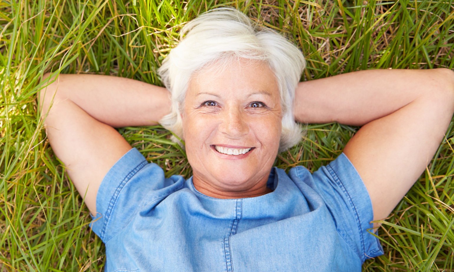 Senior woman lying on grass, resting her head on her hands, relaxing, smiling towards us