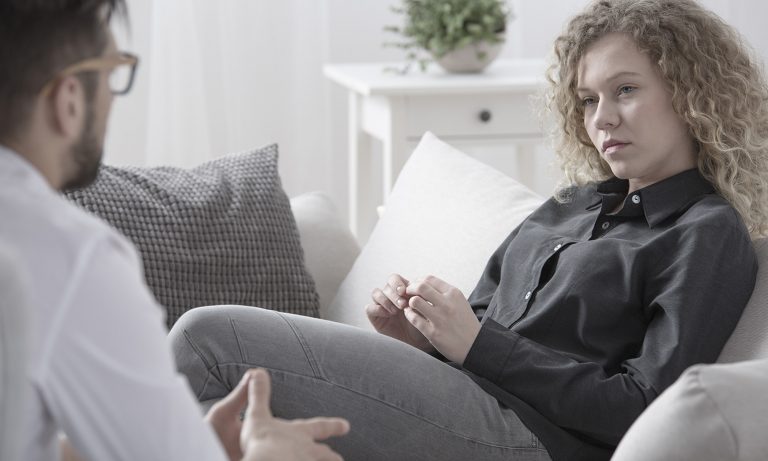 Woman in therapist's office, reclining in couch as she looks at and carefully listens to him