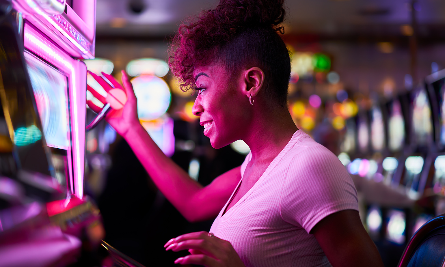 Profile view of woman smiling as she stares at slot machine, her hand on the lever, illuminated by the machine's pink light