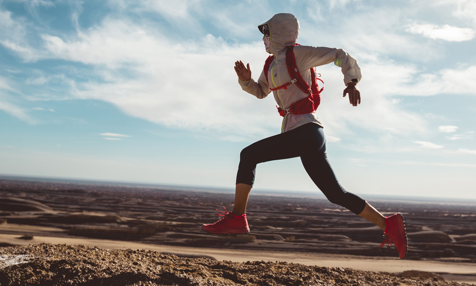 Woman, bandana over her face, runs along dirt trail in arid landscape