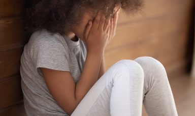 Young girl sitting on floor at edge of room, leaning against wall, hands over face, crying