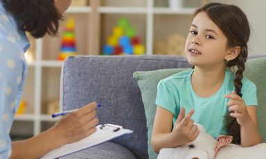 Young girl on sofa in child-oriented exam room, speaking with counselor