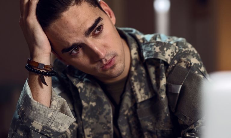 Young man wearing army fatigue jacket, leaning elbow on table, head tilted to rest against palm of his hand, his gaze distant